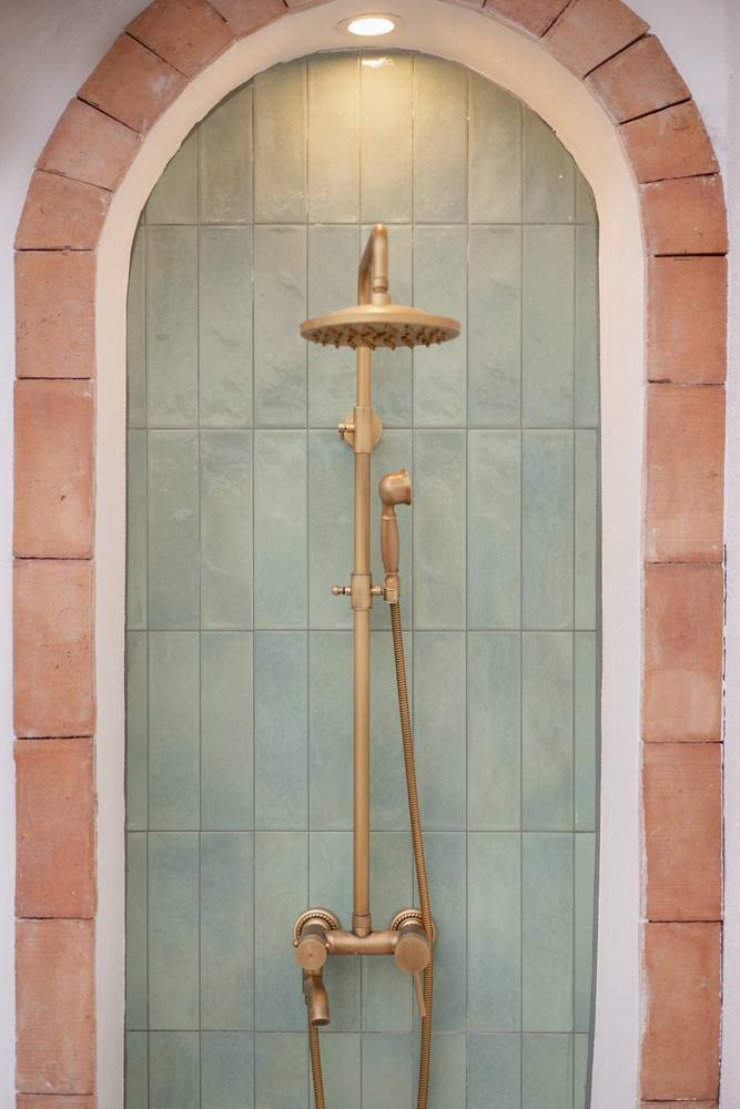 An aged-brass outdoor shower under a terracotta arch and green tiles.